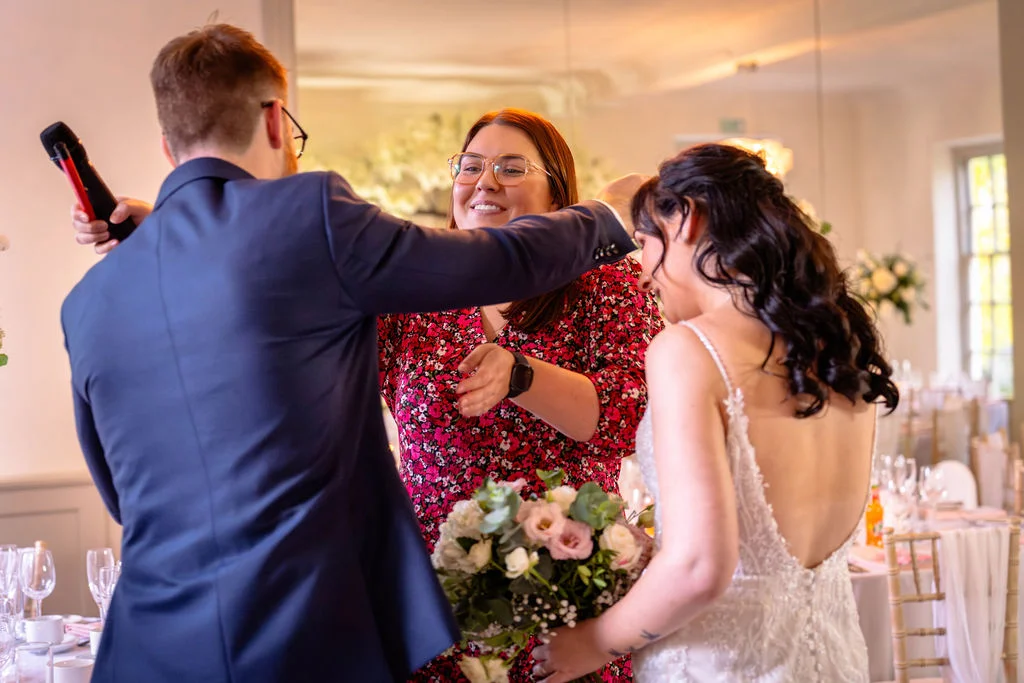 A groom in a navy suit embraces someone during the wedding speeches while the bride holds her bouquet nearby.