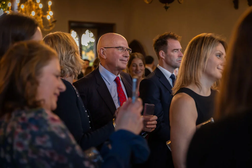 Wedding guests mingling and watching intently during a reception in a warmly lit indoor venue.