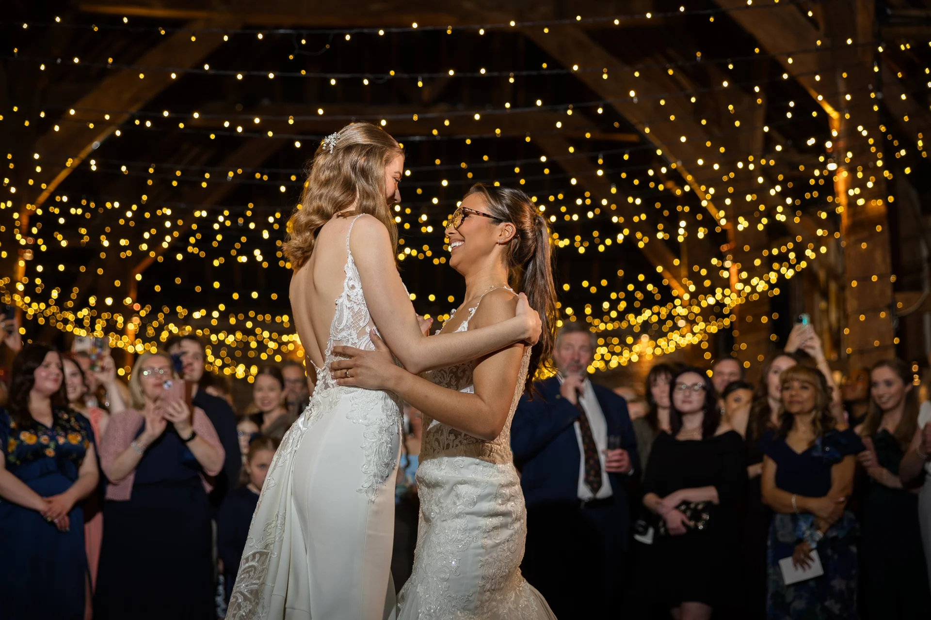 Two smiling brides share their first dance in a rustic barn illuminated by a stunning canopy of warm fairy lights overhead.