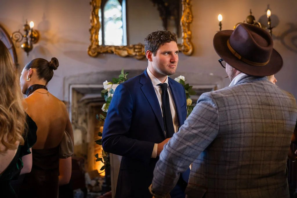 A groom in a sharp navy suit converses with a guest wearing a checked blazer and fedora inside a characterful historic venue.