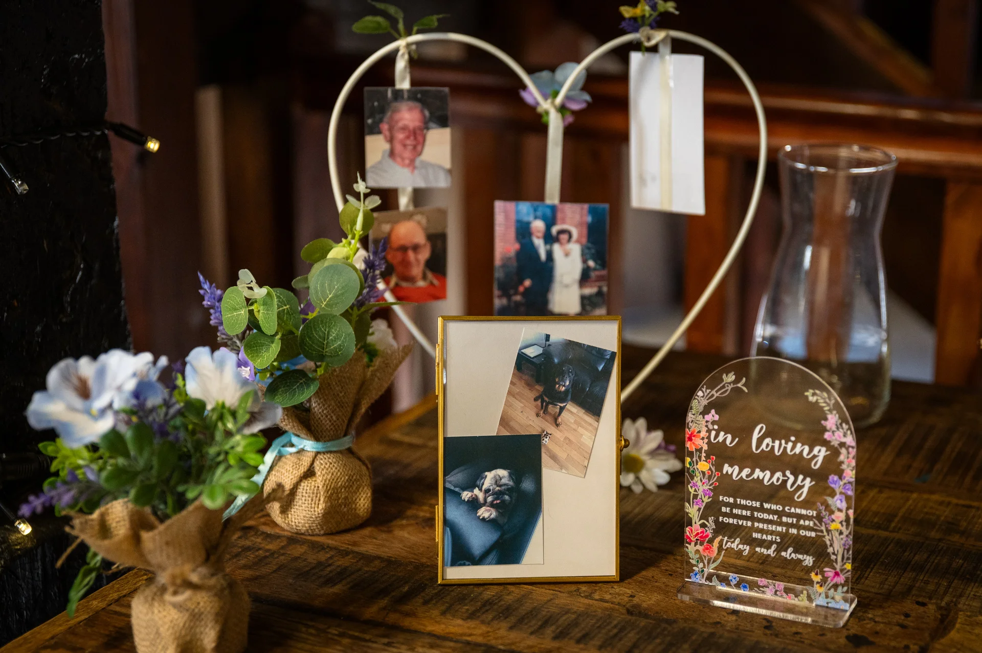 A thoughtful memory table arrangement featuring framed family photographs, an in loving memory acrylic sign, and small floral displays wrapped in hessian on a wooden surface.