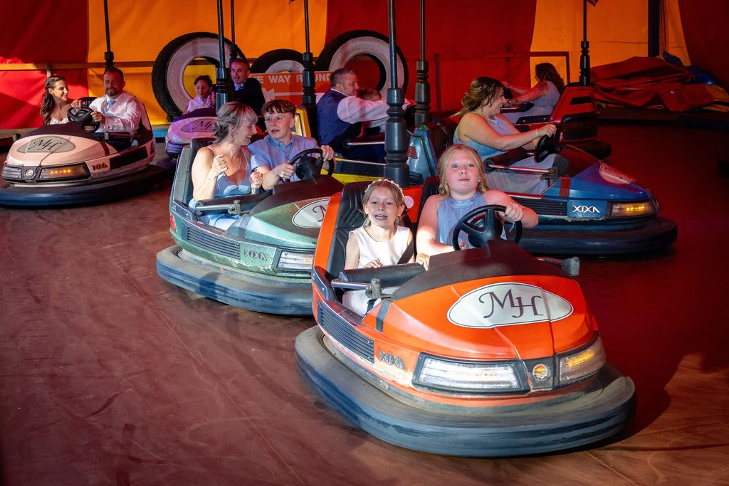 Wedding guests enjoy a fun and playful ride on fairground bumper cars inside a colourful circus tent at an evening reception.