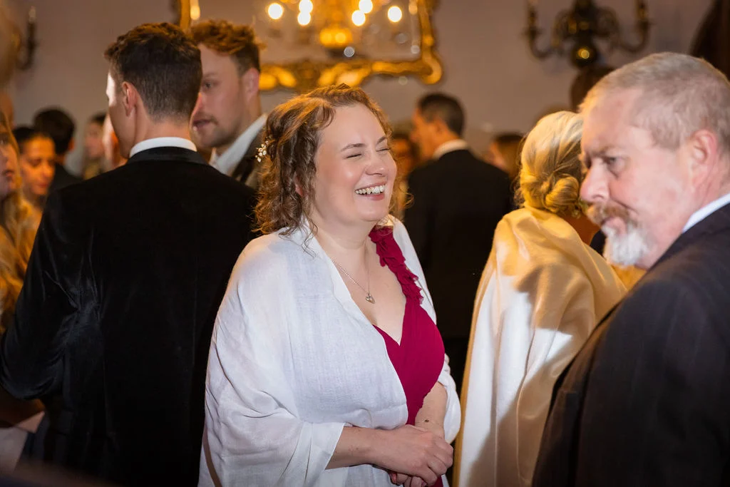 A joyful wedding guest laughs heartily while mingling with other guests inside a grand hotel reception room illuminated by a chandelier.