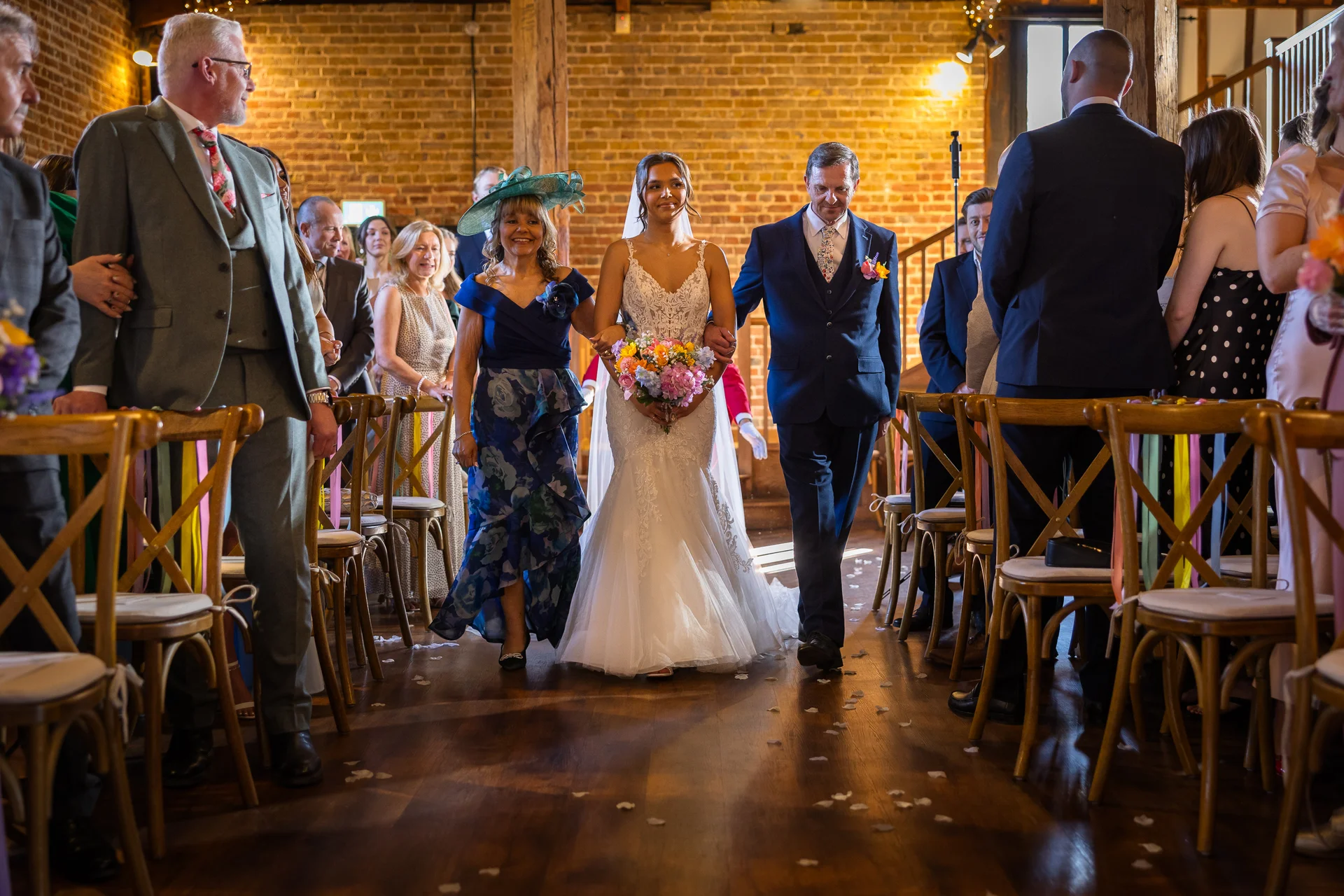The bride walks down the aisle accompanied by her parents inside a rustic brick barn venue with wooden chairs and scattered petals.