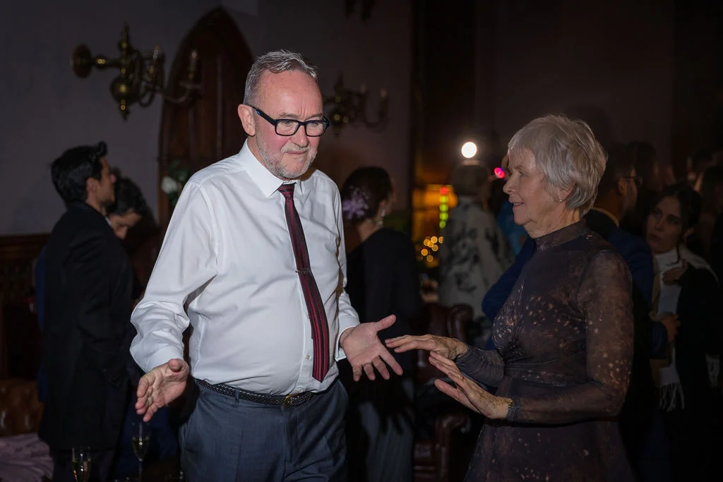An older couple enjoys dancing together at a lively evening wedding reception surrounded by guests.