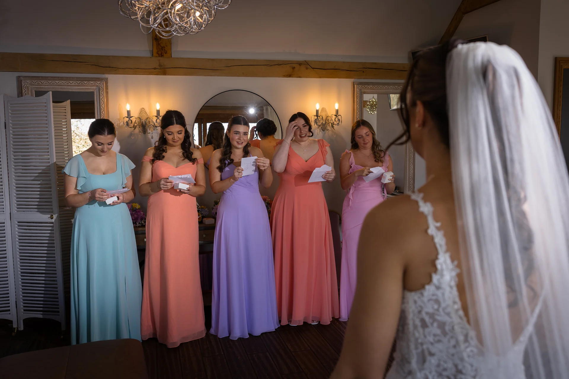 The bride in a lace gown and veil looks on as her bridesmaids in colourful pastel dresses read heartfelt letters and open gifts during their bridal prep reveal.