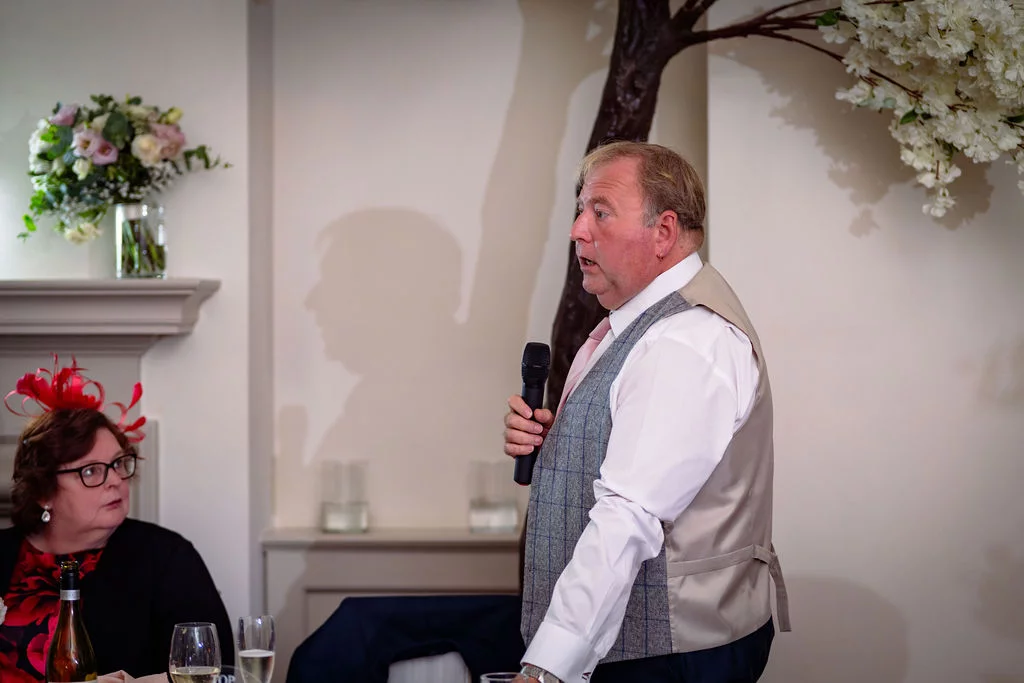 A groom delivers a speech into a microphone while wearing a tweed waistcoat, with a seated guest looking on attentively.