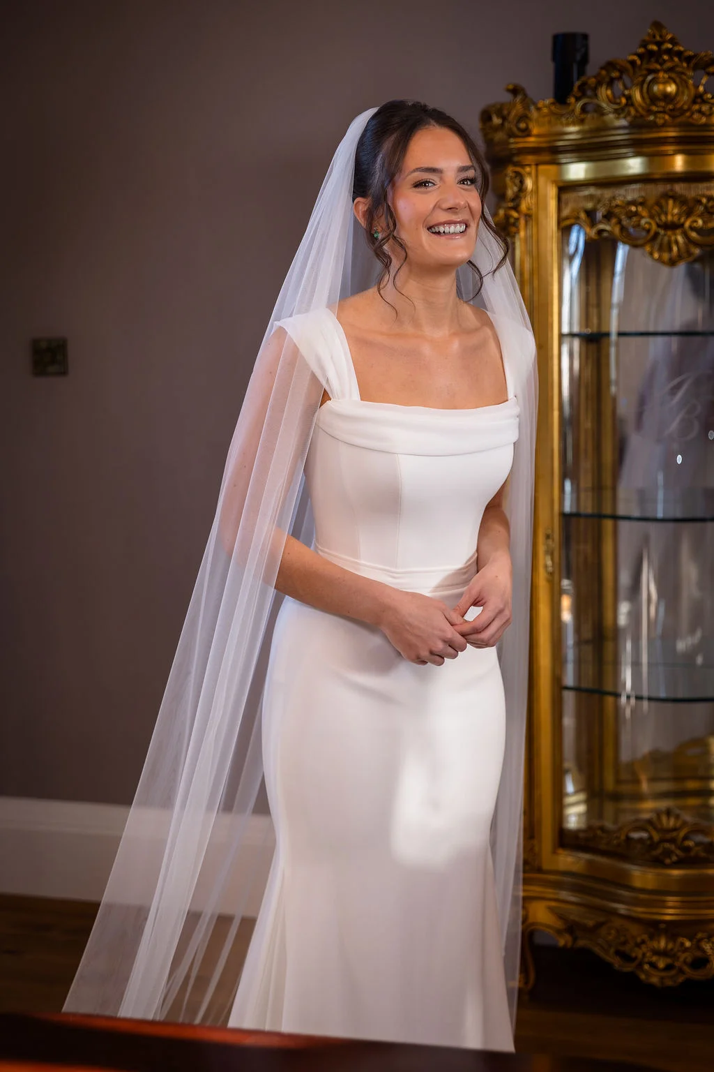 A smiling bride wearing a modern square neckline crepe fit and flare wedding dress and a long veil stands indoors next to a gold cabinet.