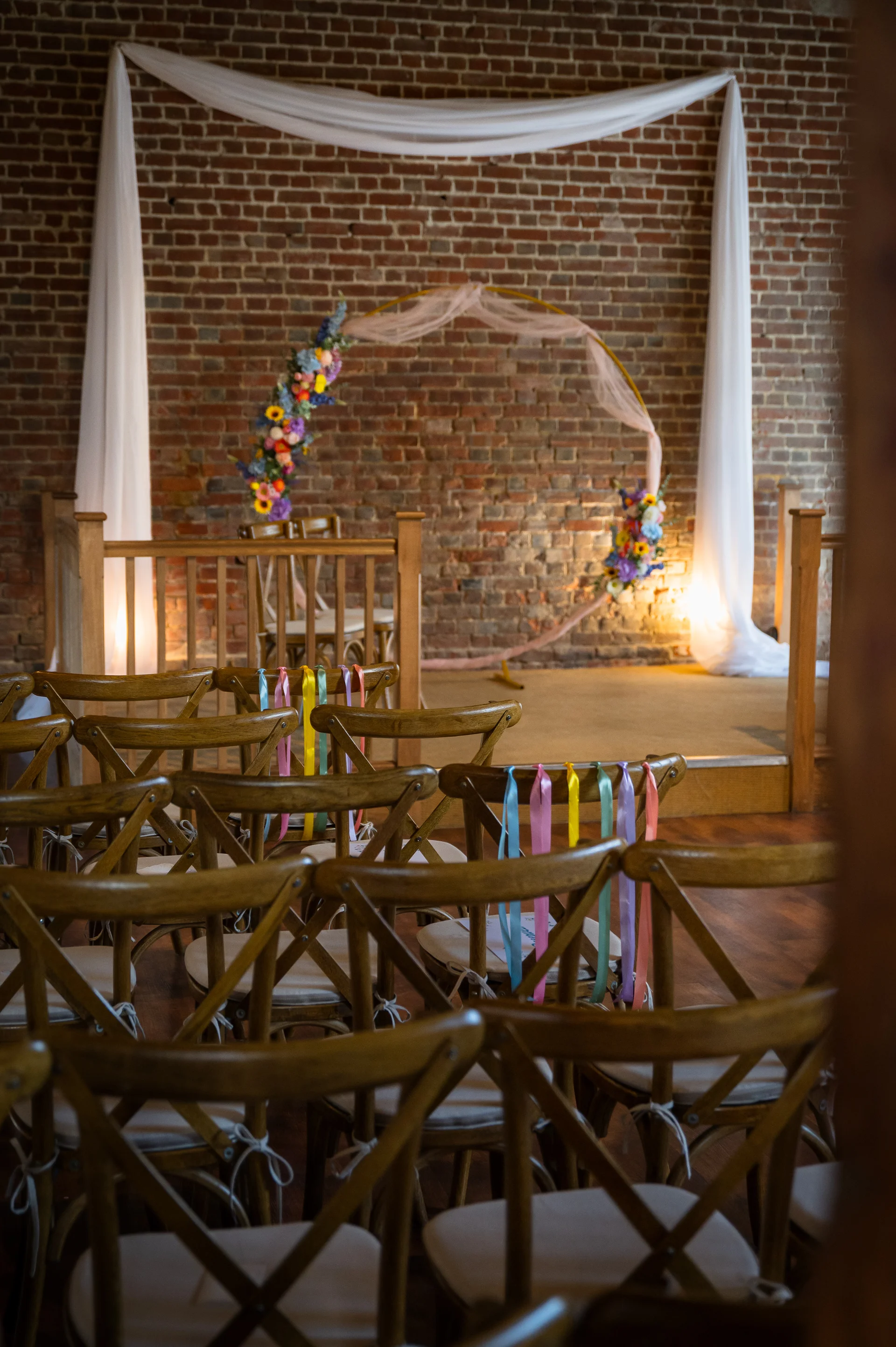 An empty wedding ceremony room featuring rustic wooden chairs adorned with colourful ribbons, facing an exposed brick wall decorated with a fabric backdrop and a floral circular arch.