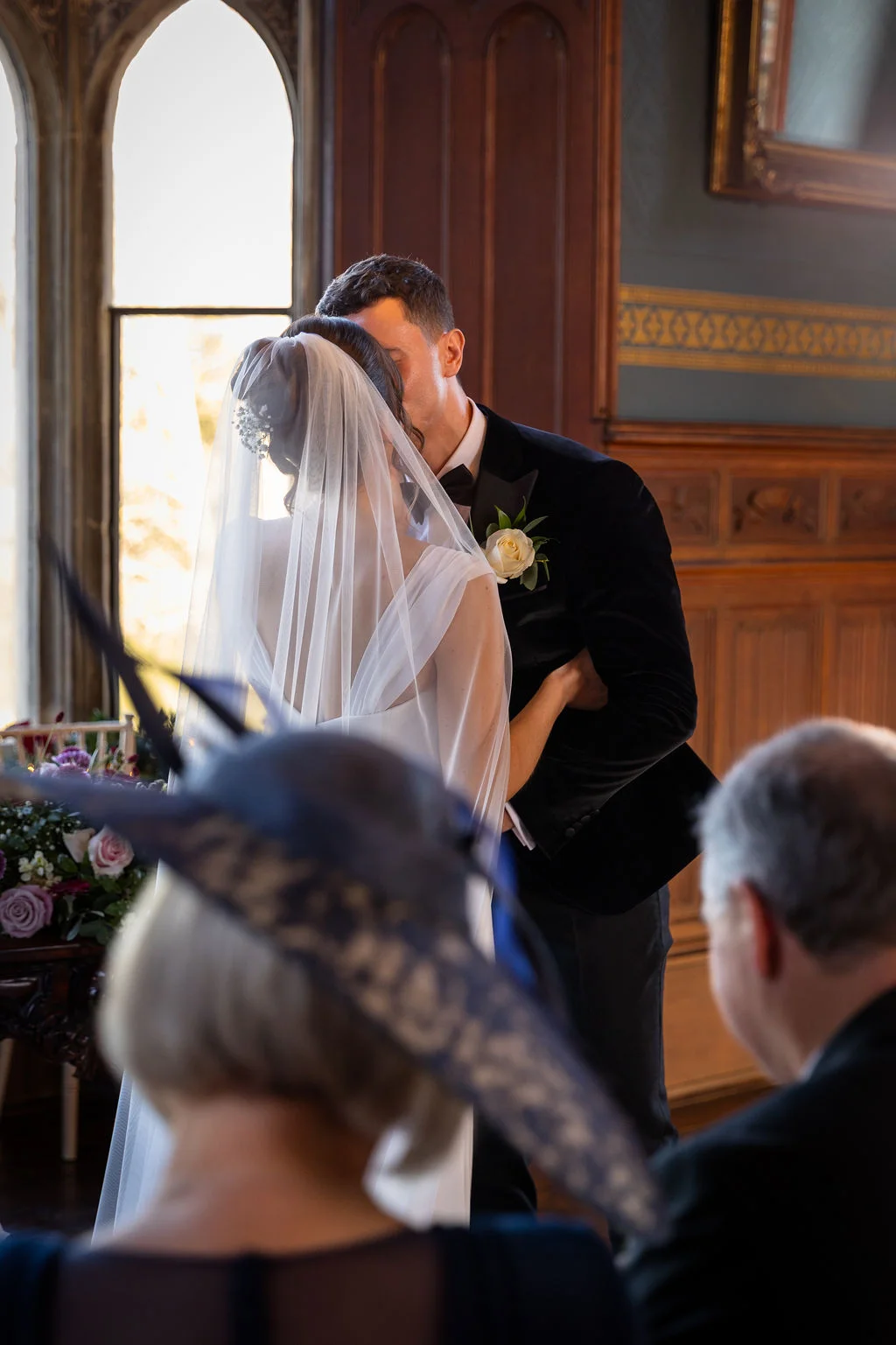 Newlyweds share their first kiss during a church ceremony, framed by guests in the foreground and warm window light.