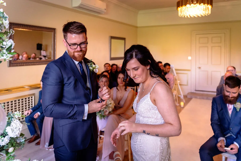 The groom places a ring on the bride's finger during their indoor civil ceremony while seated guests watch in a well-lit room.
