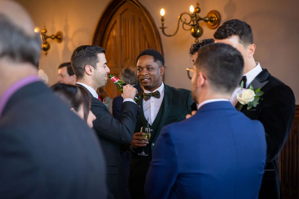 A group of wedding guests and groomsmen in suits socialise and chat together at a drinks reception indoors.