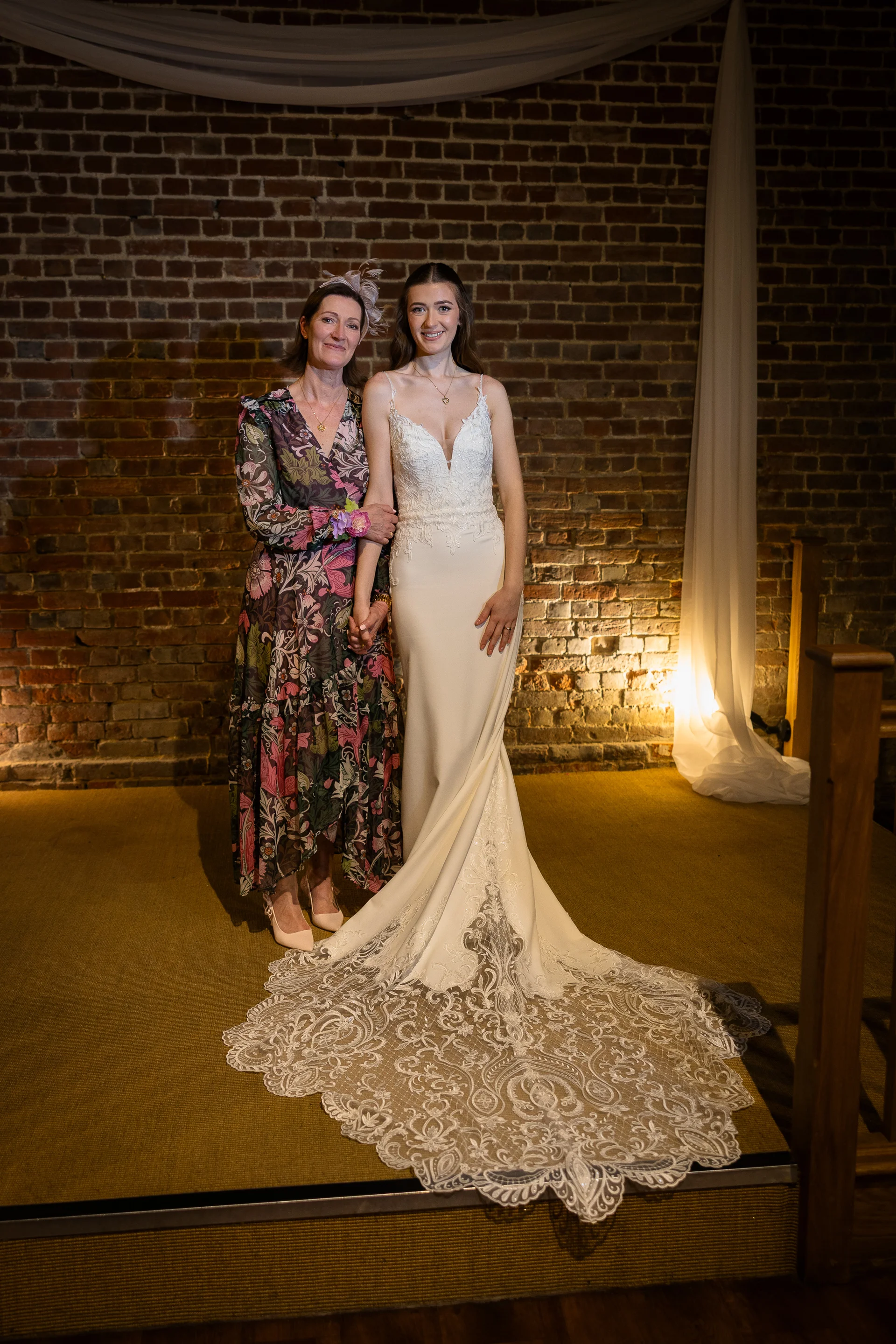A smiling bride in a fitted crepe wedding gown with a dramatic lace train poses alongside her mother in a floral dress against an exposed brick wall.