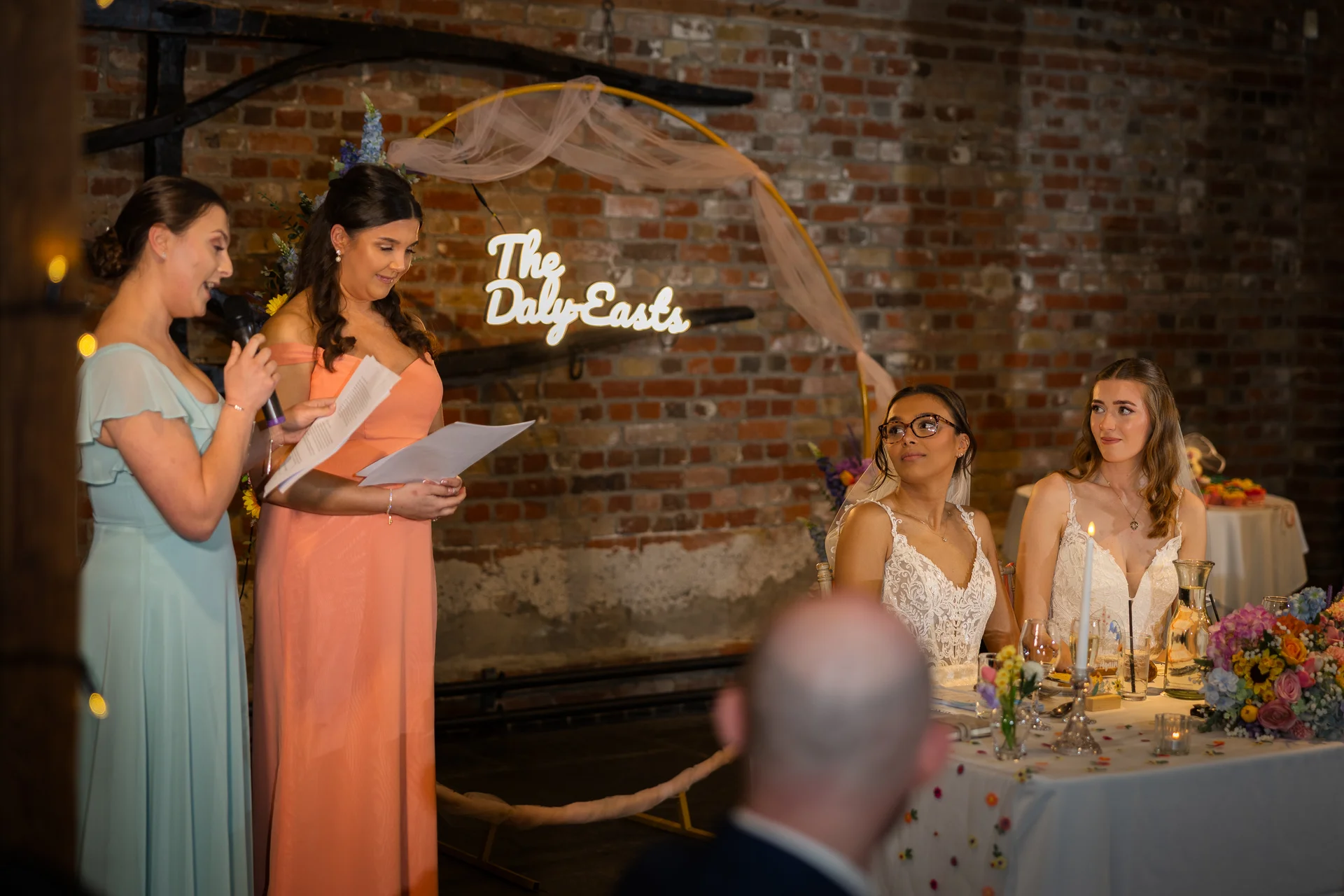 A bridesmaid delivers a speech at the top table while two brides listen attentively against an exposed brick backdrop featuring a custom neon sign.
