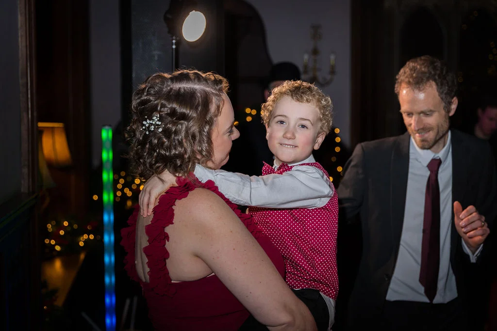 A woman in a red dress holds a young boy in a polka dot waistcoat during a lively evening wedding reception with dancing guests.