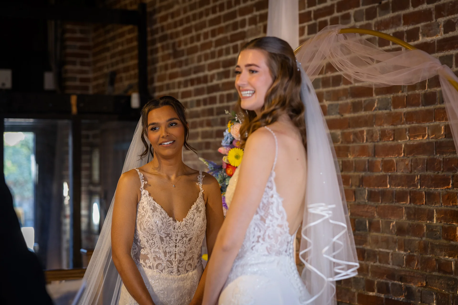 Two smiling brides stand side by side wearing matching lace wedding dresses and veils in front of an exposed brick wall during their ceremony.