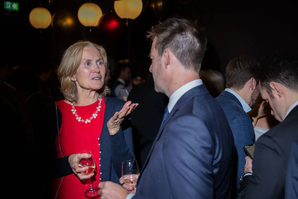 A mature female guest in a red dress speaks animatedly while holding a wine glass during a lively evening drinks reception.