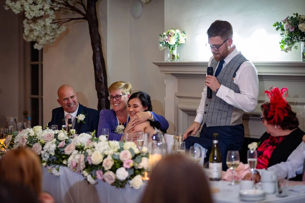 A groom delivers an emotional wedding speech while a family member hugs the blushing bride seated at the flower-adorned top table.