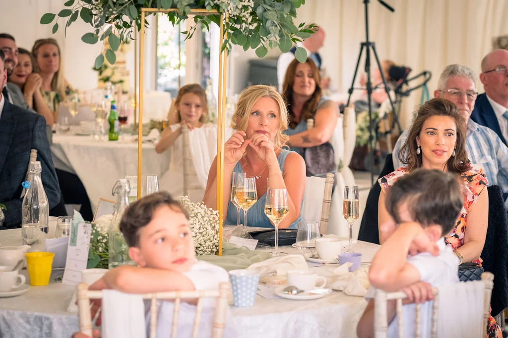 Guests are seated at round tables during the wedding breakfast, listening attentively while listening to speeches.