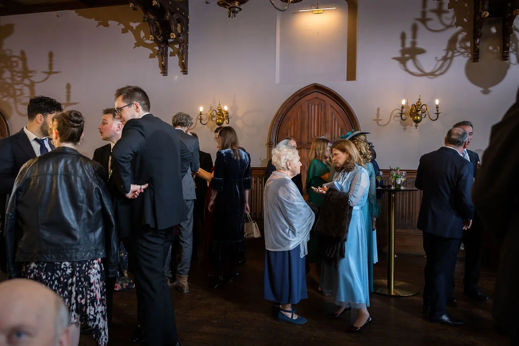 Wedding guests mingle and converse in a grand, wood-panelled historic hall during the drinks reception.