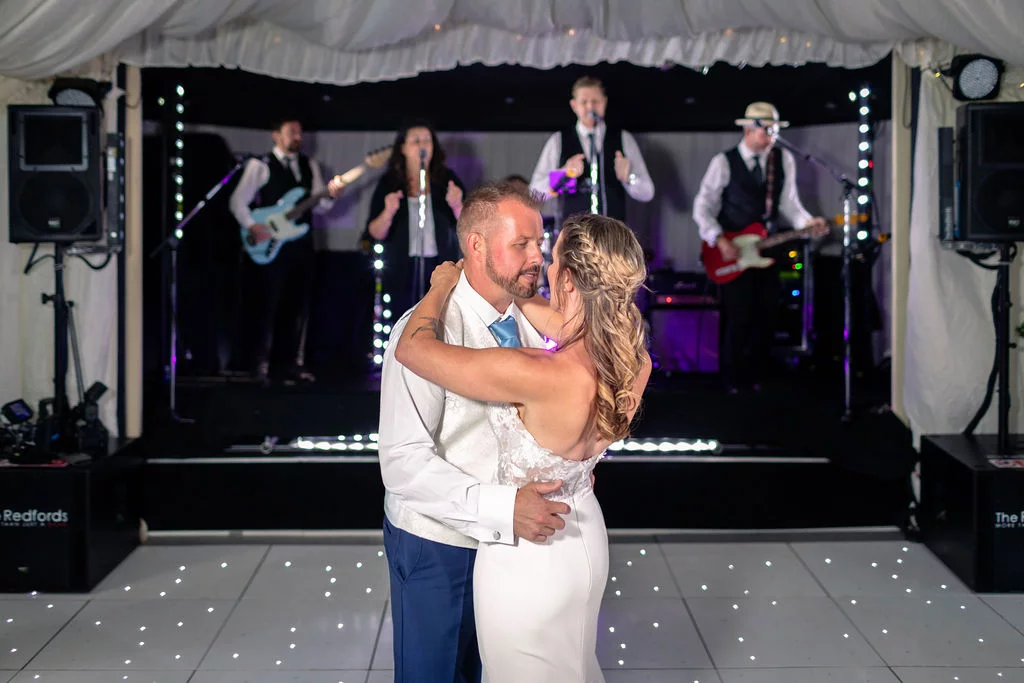 The newlywed couple share their first dance on a starlit dance floor inside a marquee while a live band performs on stage behind them.
