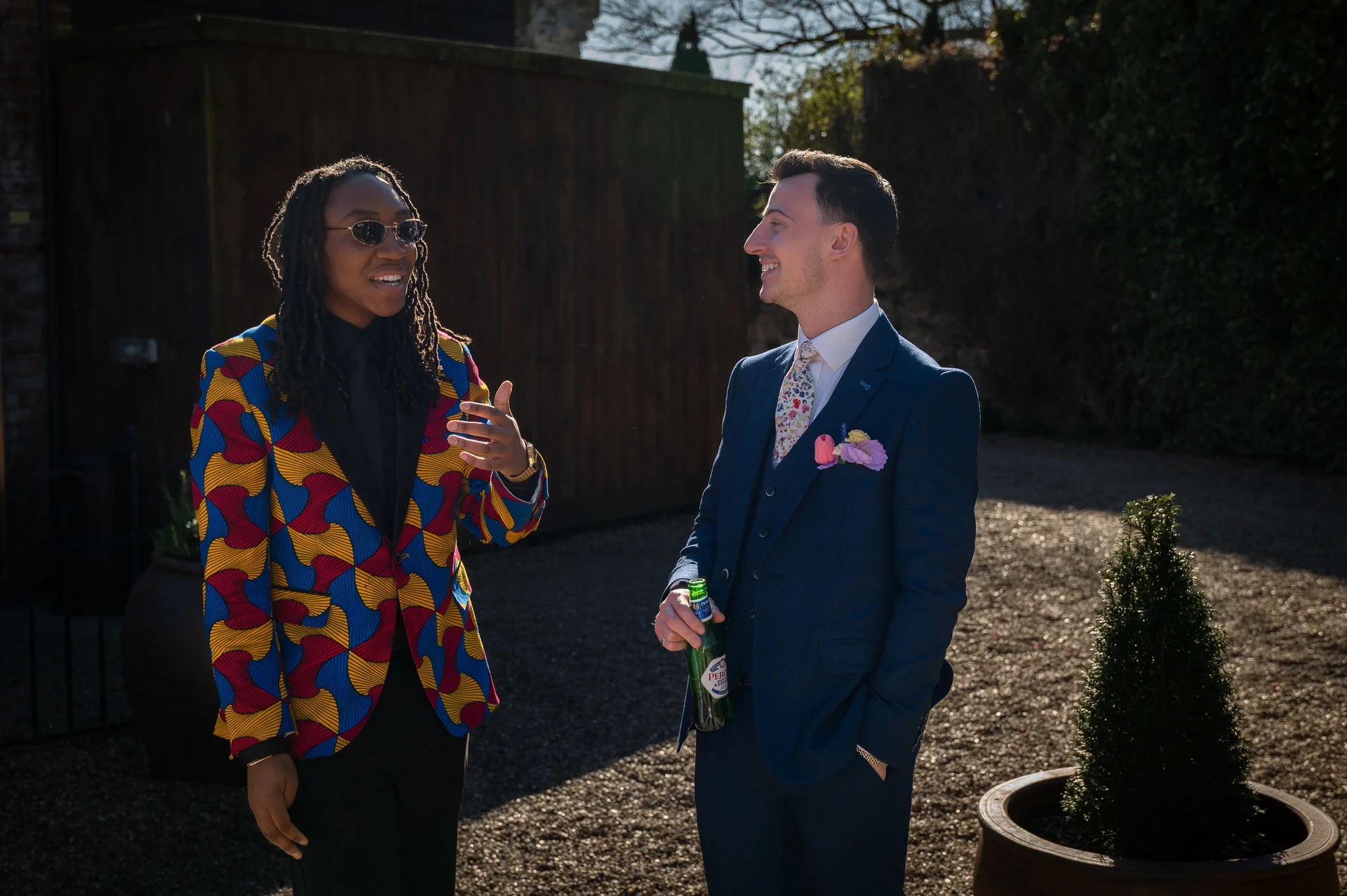 Two grooms in stylish wedding attire laugh together outside in bright sunshine, with one wearing a vibrant patterned jacket and sunglasses while holding a beer.