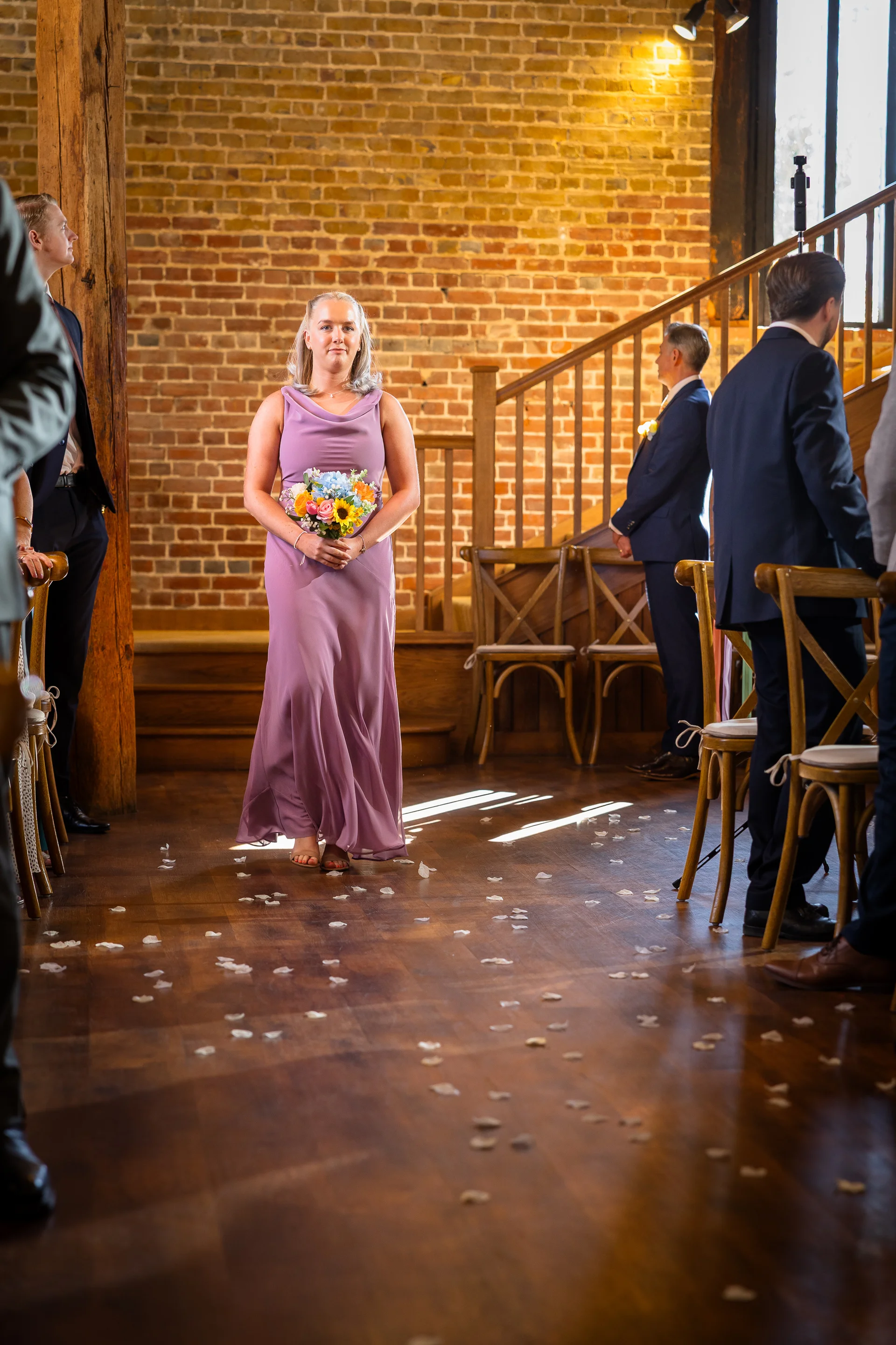 A bridesmaid wearing a pink dress walks down the aisle strewn with petals inside a rustic brick barn.