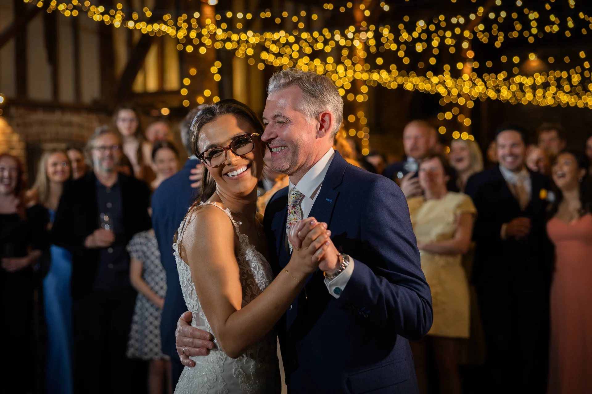 A smiling bride wearing glasses and a lace gown shares a joyful first dance with her groom under a glowing canopy of fairy lights inside a rustic barn.