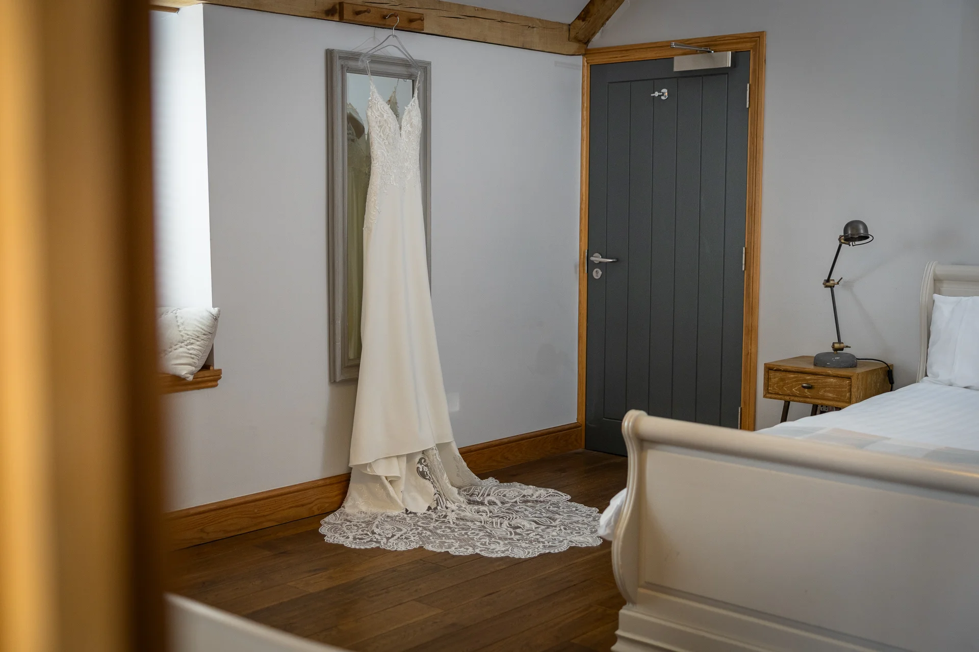 An elegant lace wedding dress hangs gracefully from a mirror on a bedroom wall, showcasing its intricate train pooling on the wooden floor.