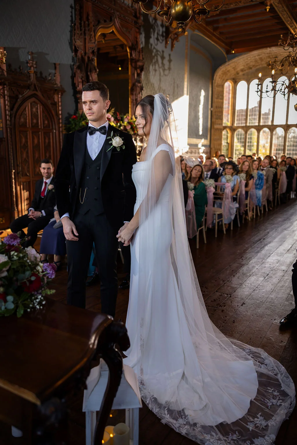 A newly married couple stand at the front of a grand, wood-panelled hall during their wedding ceremony, bathed in warm light from tall arched windows.