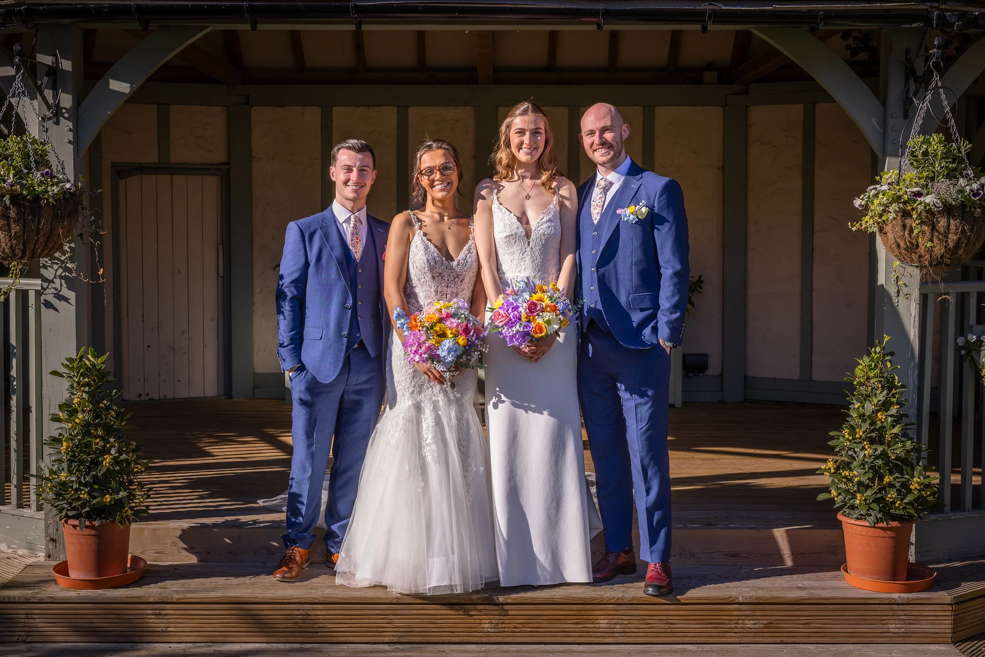 A happy two-bride couple stands proudly with their wedding party outside on a wooden deck in bright sunshine, holding colourful floral bouquets.