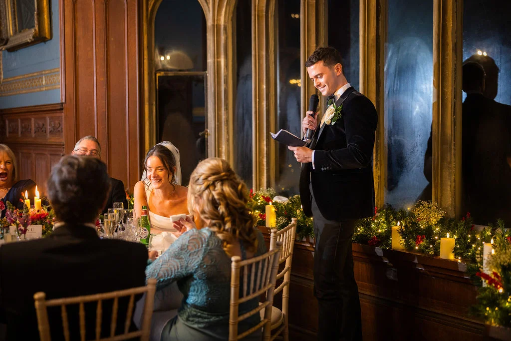 The groom delivers an entertaining speech during the wedding breakfast while the bride laughs heartily seated at the candlelit top table.