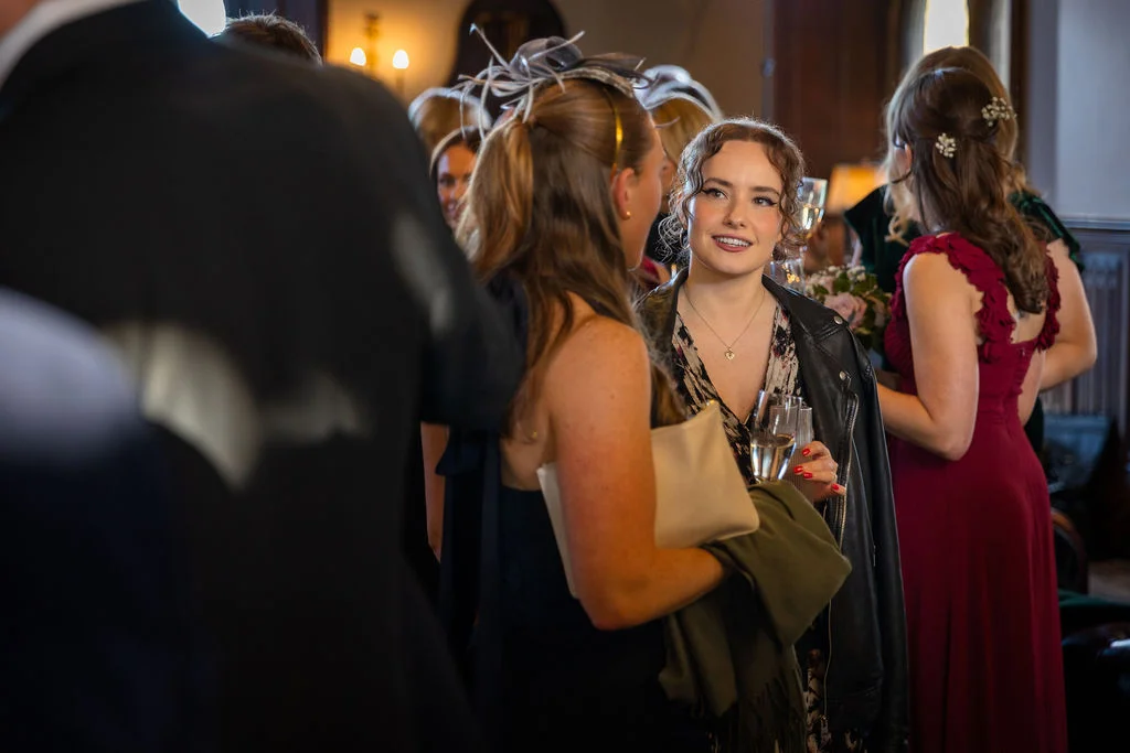 Wedding guests mingle happily during the drinks reception, holding glasses of champagne while dressed in stylish cocktail attire and fascinators.