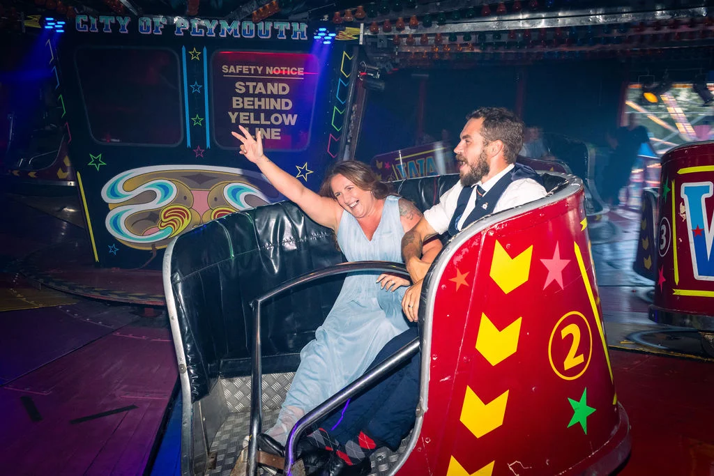 A joyful couple laughs and raises their hands in the air while riding a vibrant red fairground car at their evening wedding reception.
