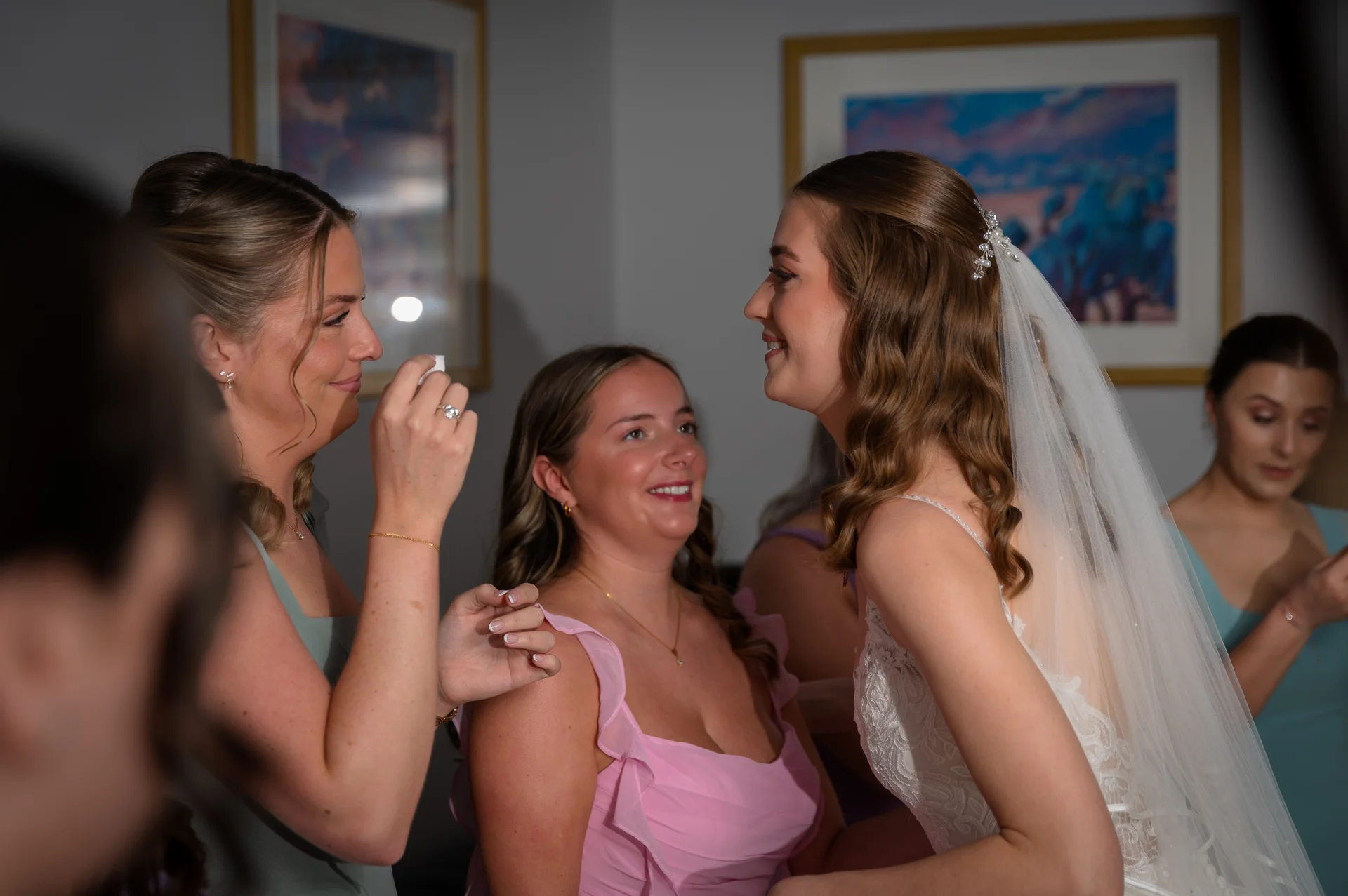 A smiling bride in a lace wedding dress and veil shares a joyful moment with her bridesmaids during morning bridal preparations.