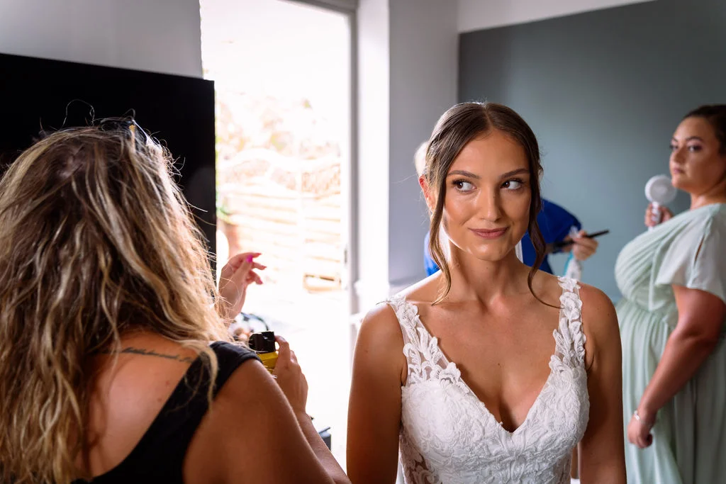 A bride in a lace v-neck wedding dress smiles during her final makeup touch-ups as she looks into a mirror on the morning of her wedding.