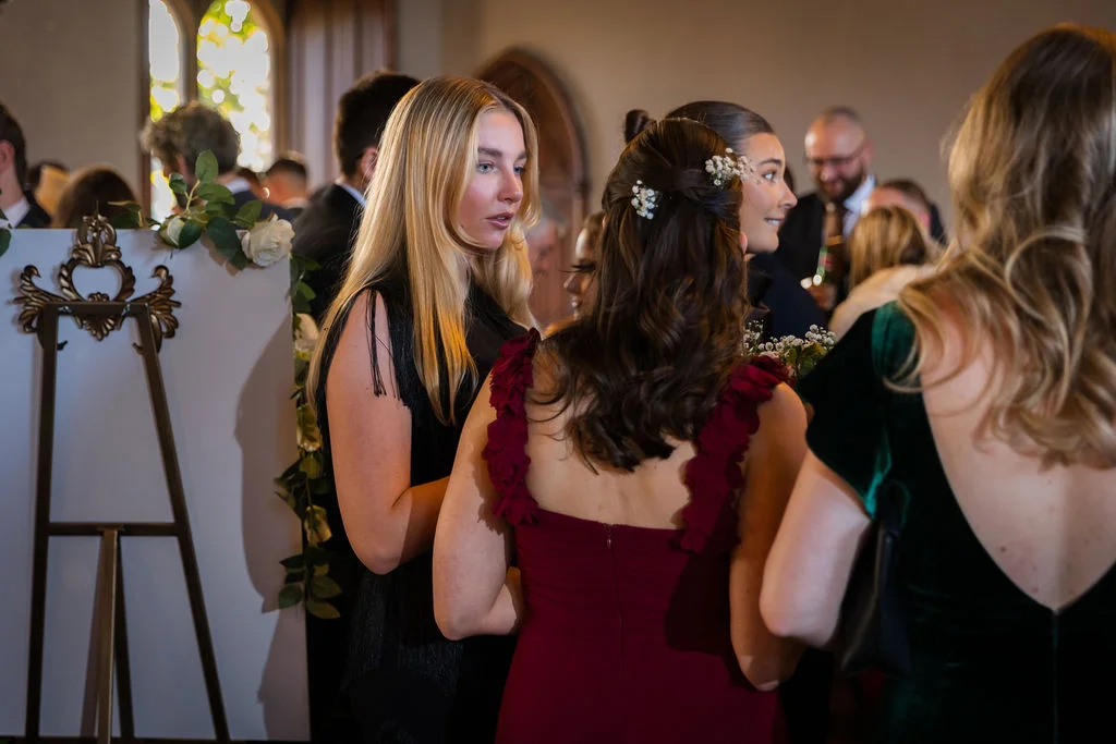 Wedding guests mingling indoors, including a woman with long blonde hair talking and another wearing a burgundy dress with ruffled straps.