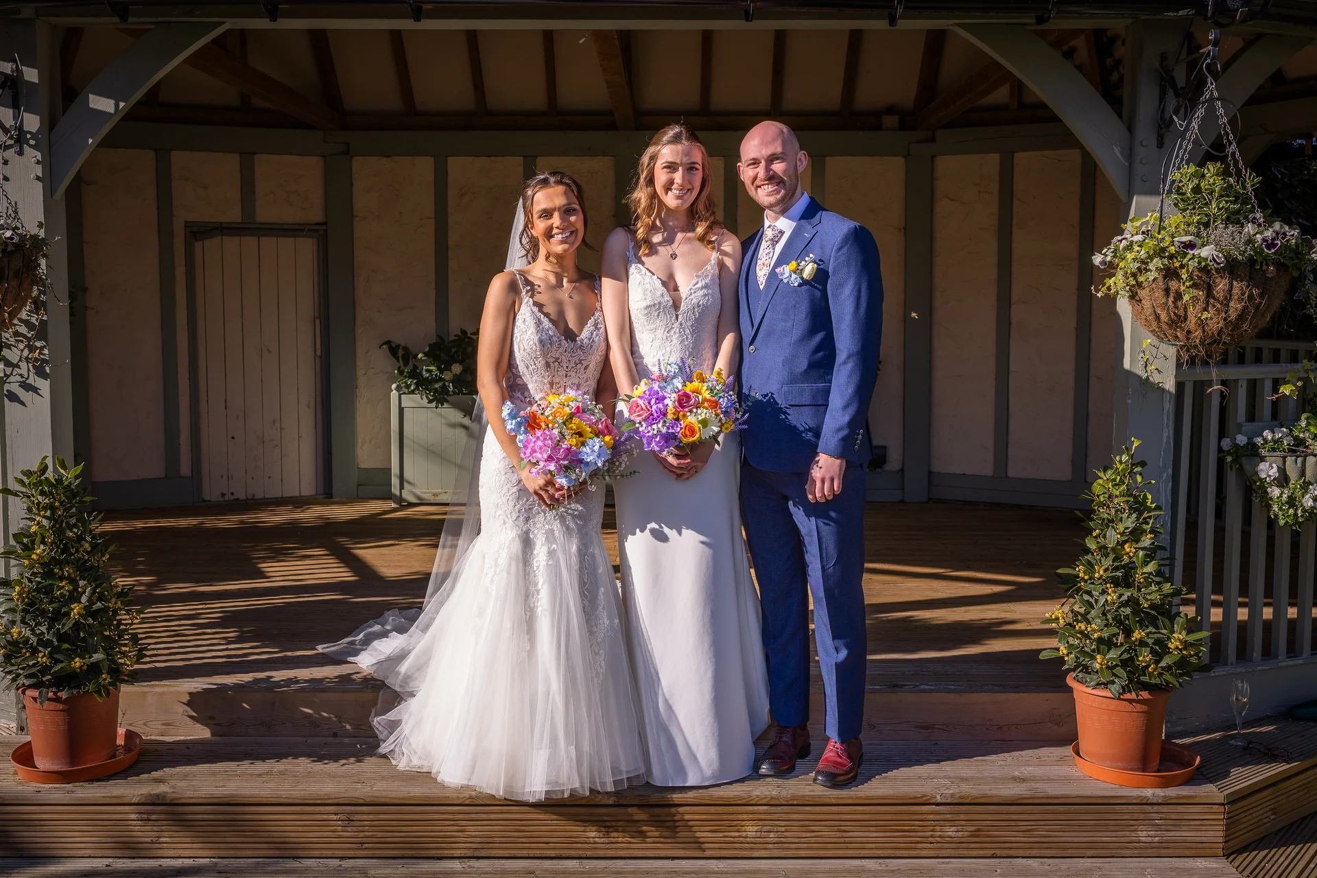 A smiling same-sex couple and a groom pose together on a wooden pavilion deck holding colourful vibrant wedding bouquets in bright sunlight.