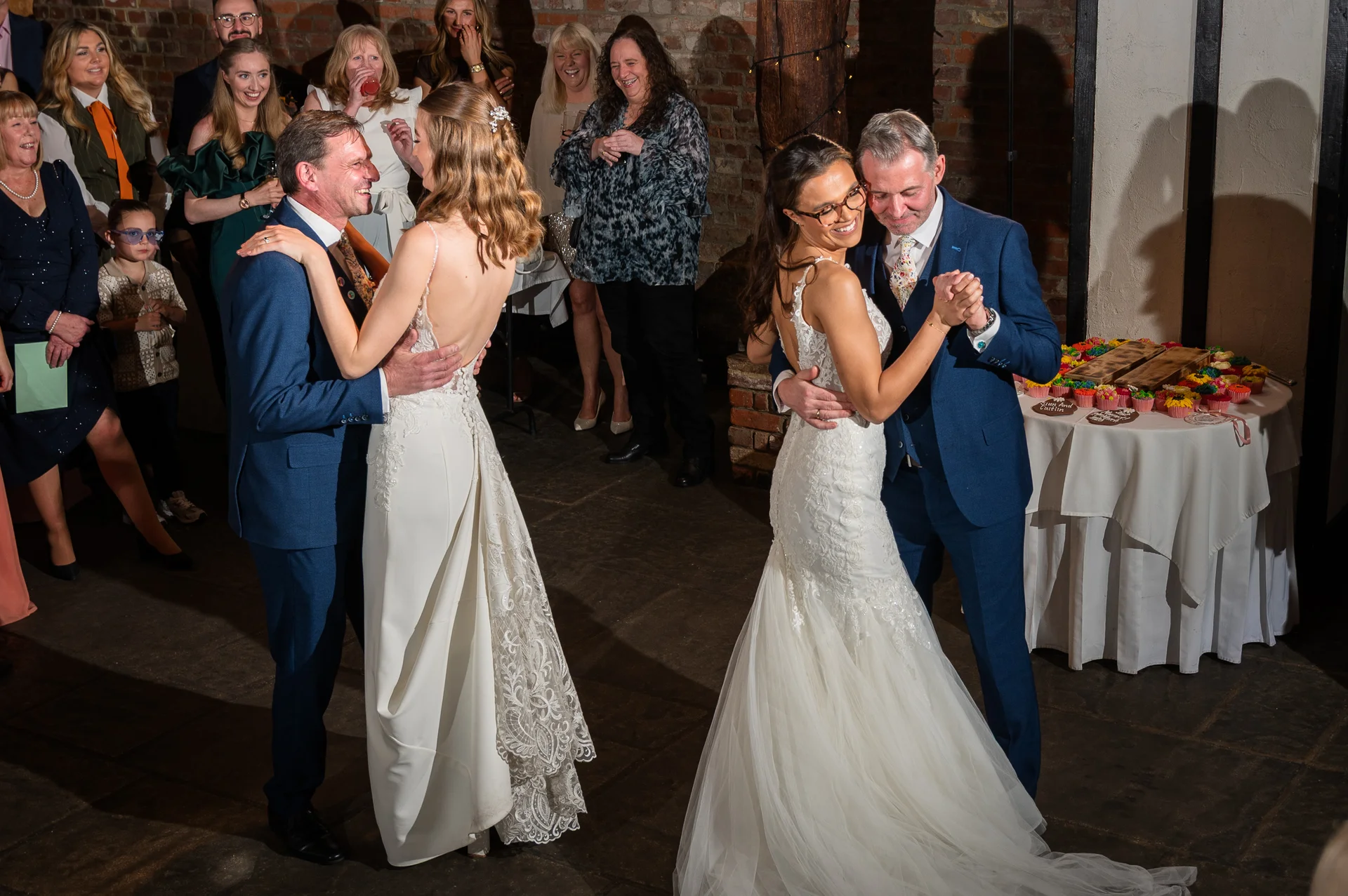 Two brides in stunning lace wedding gowns share their first dance with their partners in blue suits, surrounded by joyful guests in a rustic venue.
