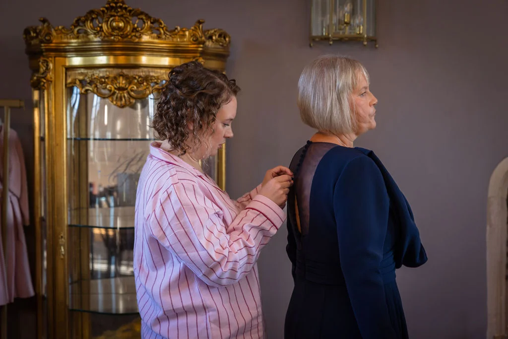 A woman in pink striped pyjamas carefully fastens the buttons on a dark blue dress worn by a mature woman during bridal preparations.