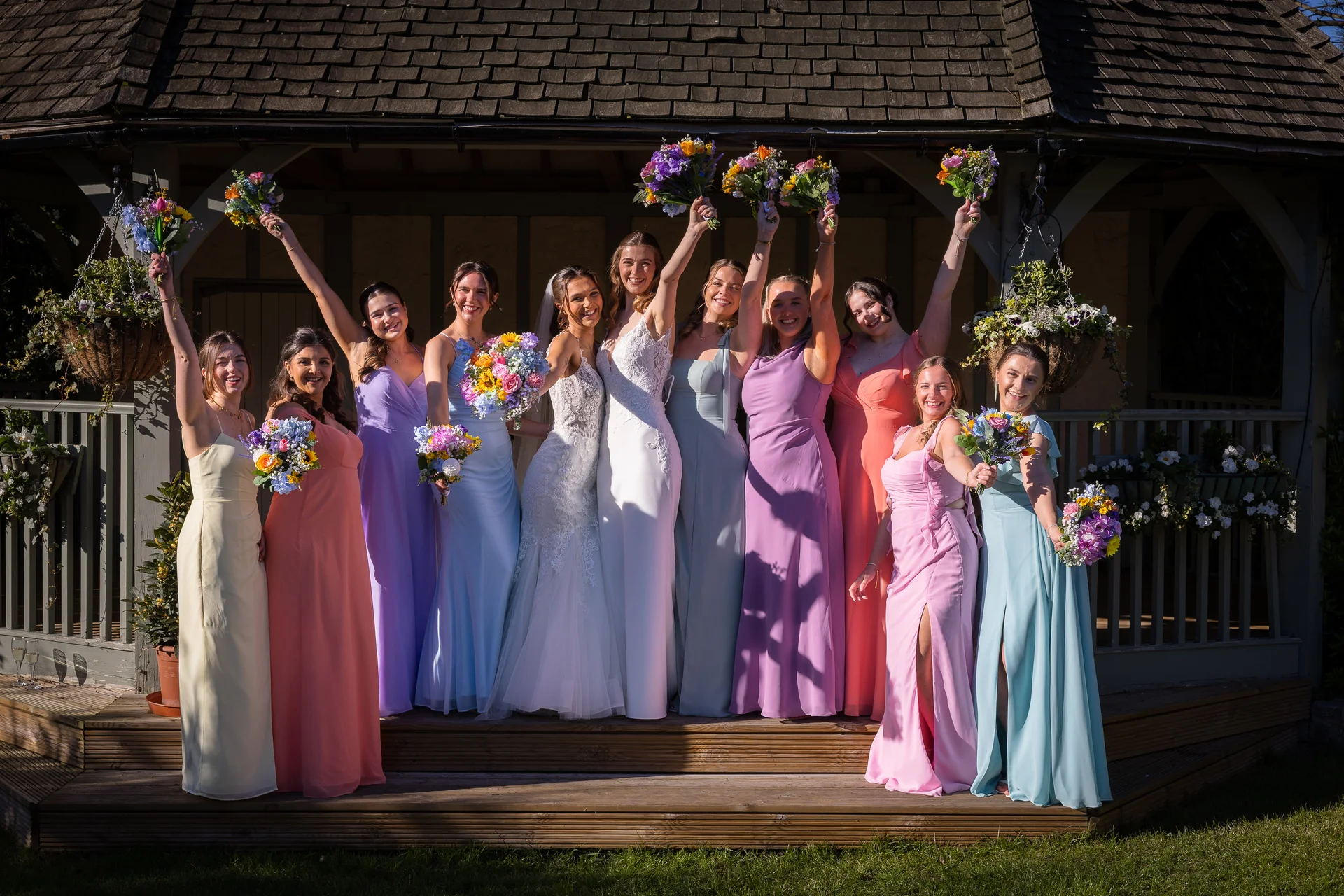 A radiant bride and her large bridal party of mismatched pastel-clad bridesmaids raise their colourful bouquets in celebration outside a rustic wooden gazebo.