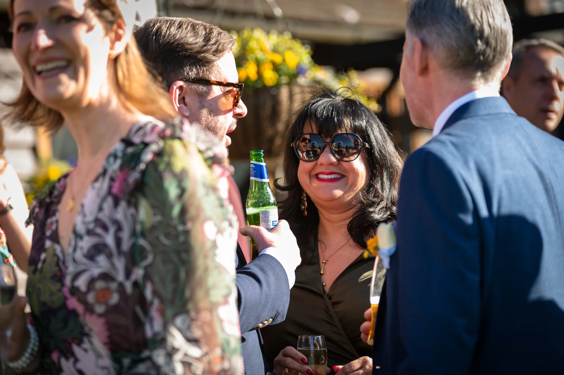 Wedding guests laugh and mingle together outside during a sunny drinks reception, holding drinks in the warm afternoon sunlight.