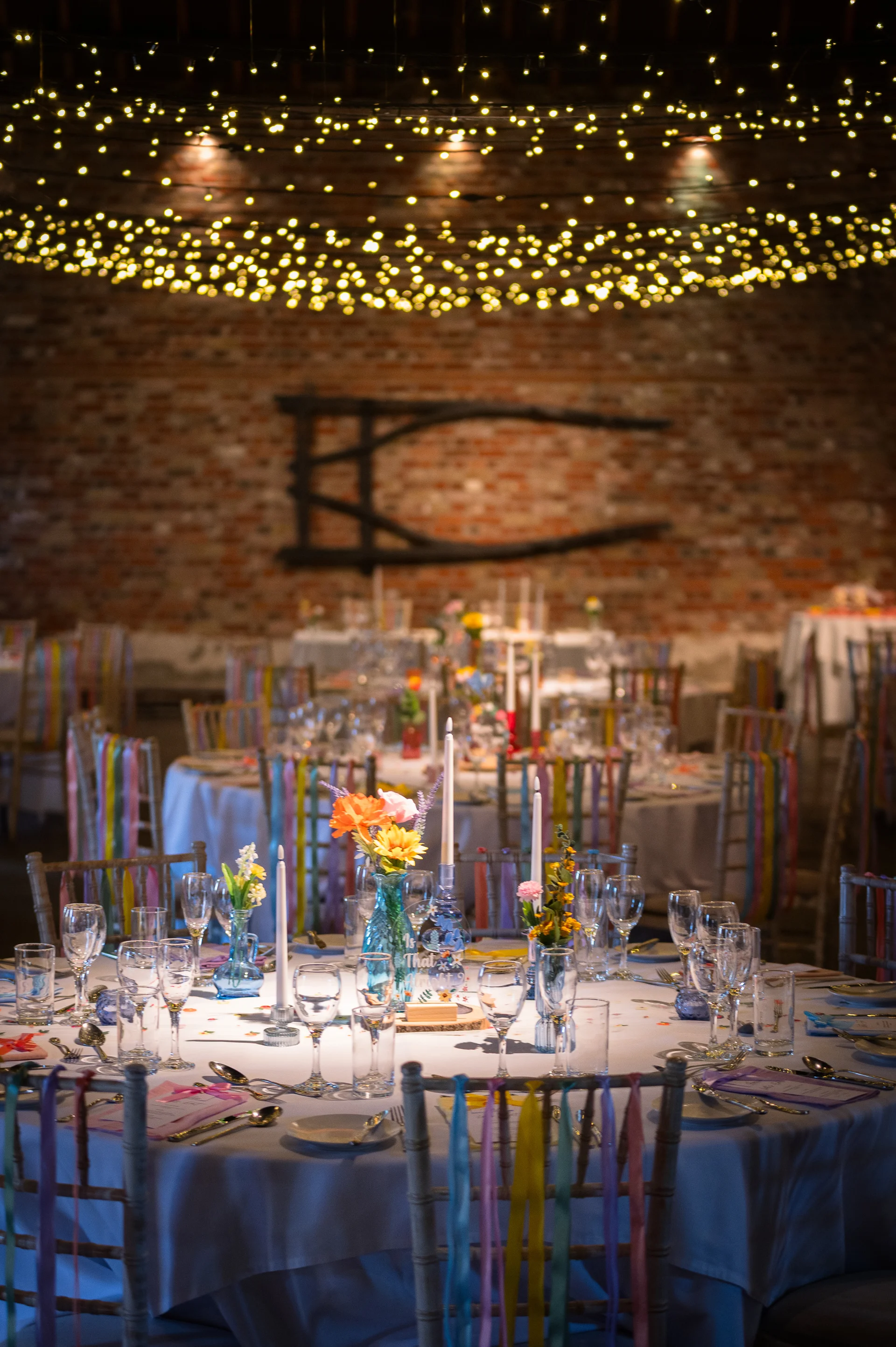 A beautifully styled wedding reception in a rustic barn featuring round tables with white linens, colourful ribbon-adorned chairs, vibrant floral bud vases, tapered candles, and an extensive fairy light canopy overhead against an exposed brick wall.