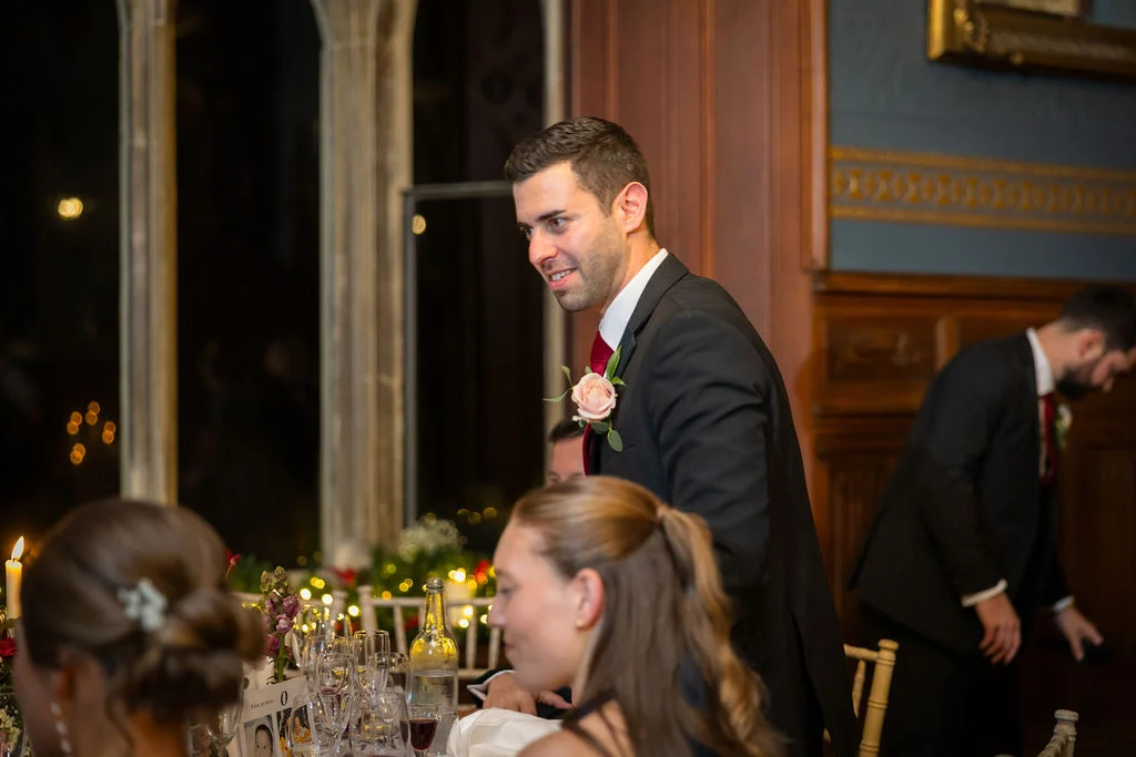 A groom smiling during his wedding speech inside a wood-panelled reception room, wearing a black suit with a pink rose boutonniere.