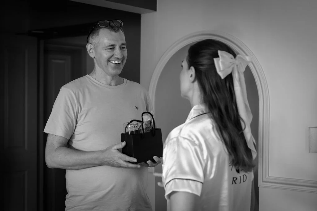 A joyful father smiling broadly while presenting a gift bag to his daughter who is wearing a matching satin pyjama set and a hair bow during bridal preparations.