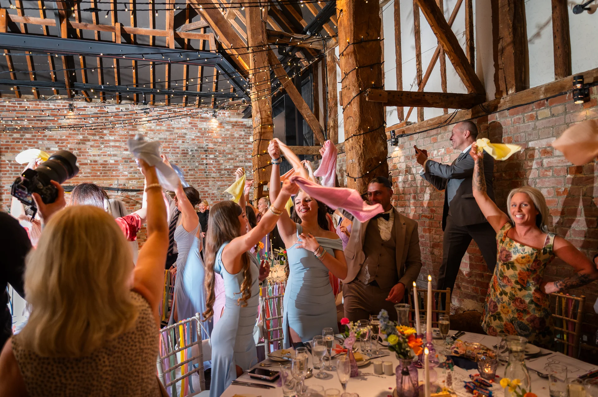 Wedding guests enthusiastically wave napkins in the air during a lively reception inside a rustic brick and timber barn.
