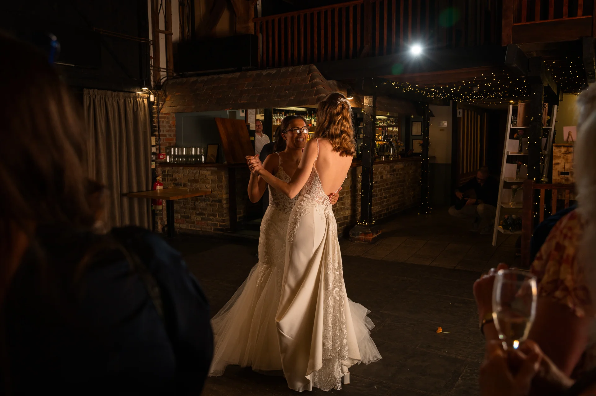 Two brides share a joyful first dance together in a rustic barn venue illuminated by warm fairy lights.