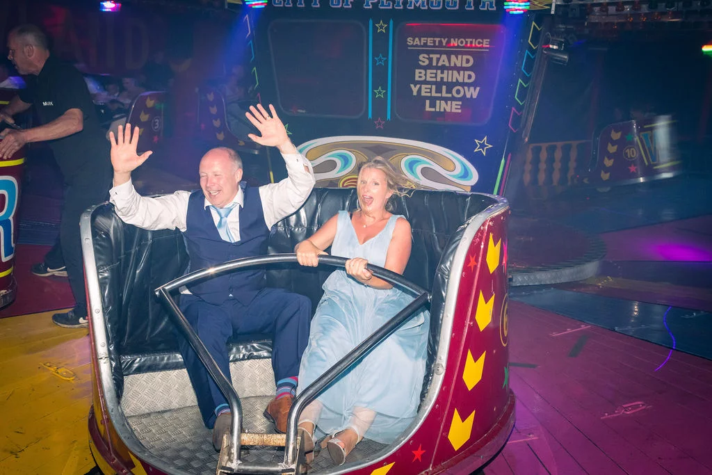 A newlywed couple laughing and waving while riding a fairground bumper car together during their evening wedding reception.