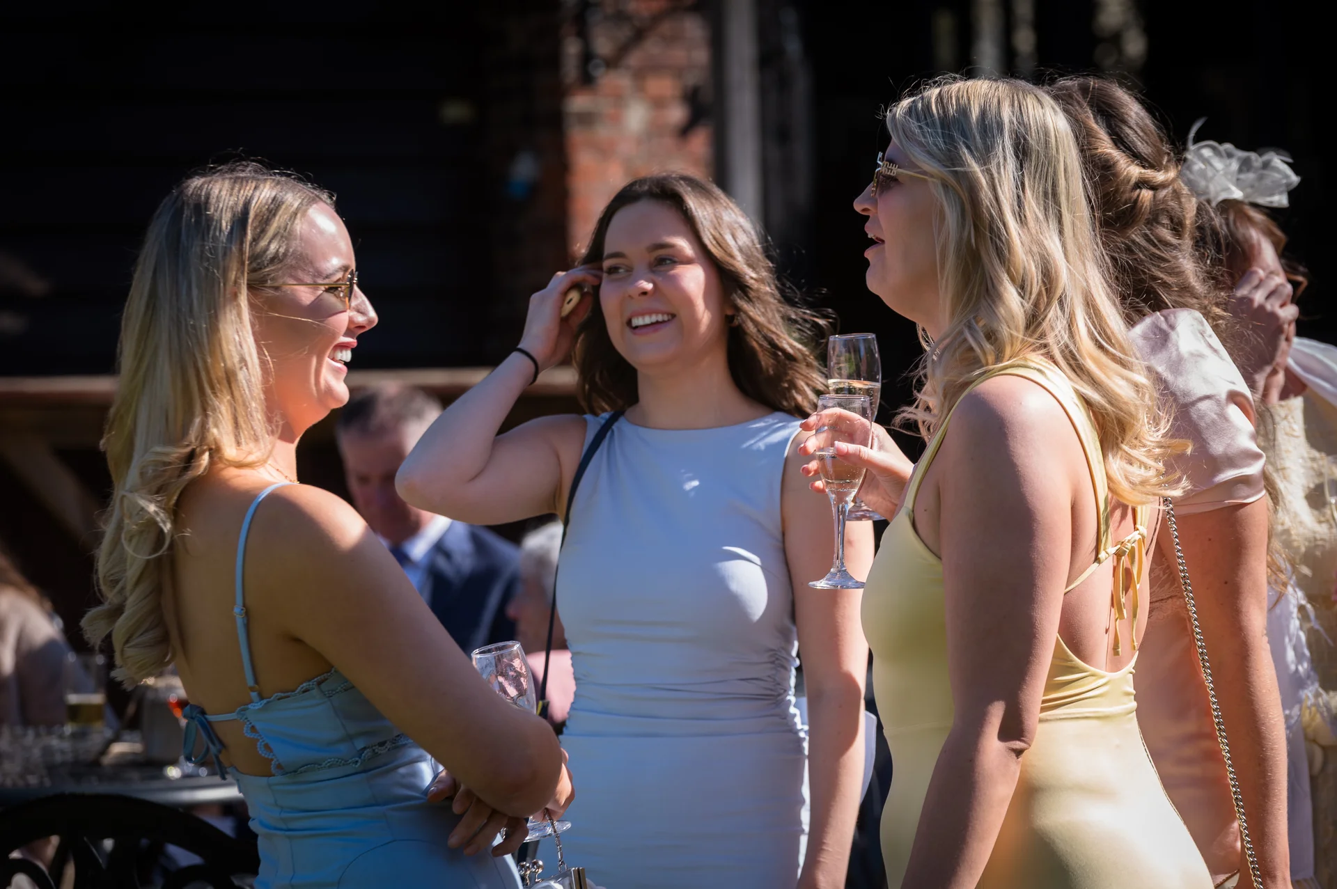 A group of female wedding guests laugh and chat together outdoors during the drinks reception, holding champagne flutes in the bright sunshine.