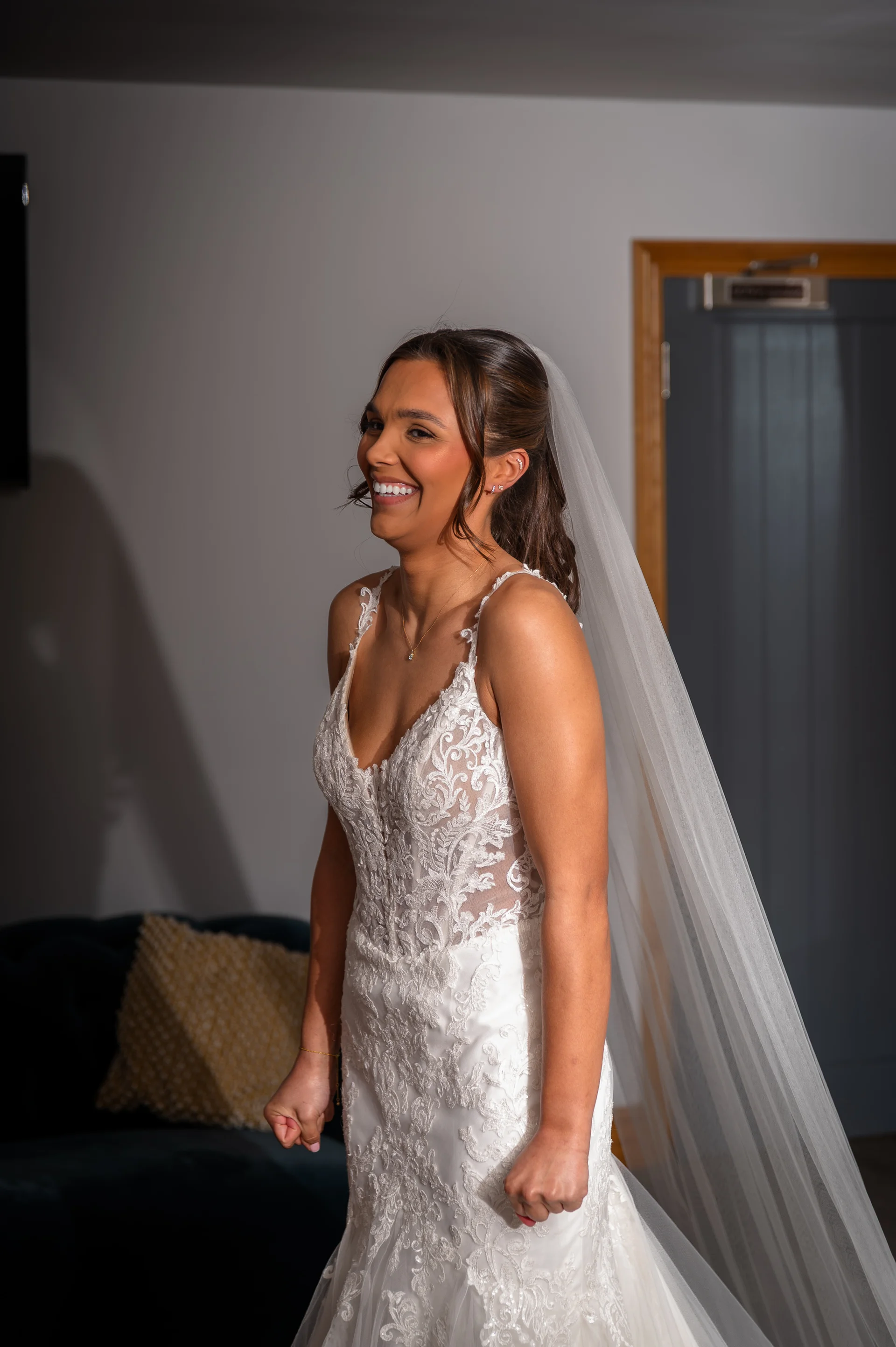 A joyful bride smiles broadly while wearing a lace appliquéd gown with spaghetti straps and a flowing veil during morning preparations.